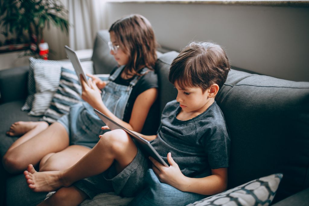 Two children sitting on a sofa using tablets indoors, focused on the screens.